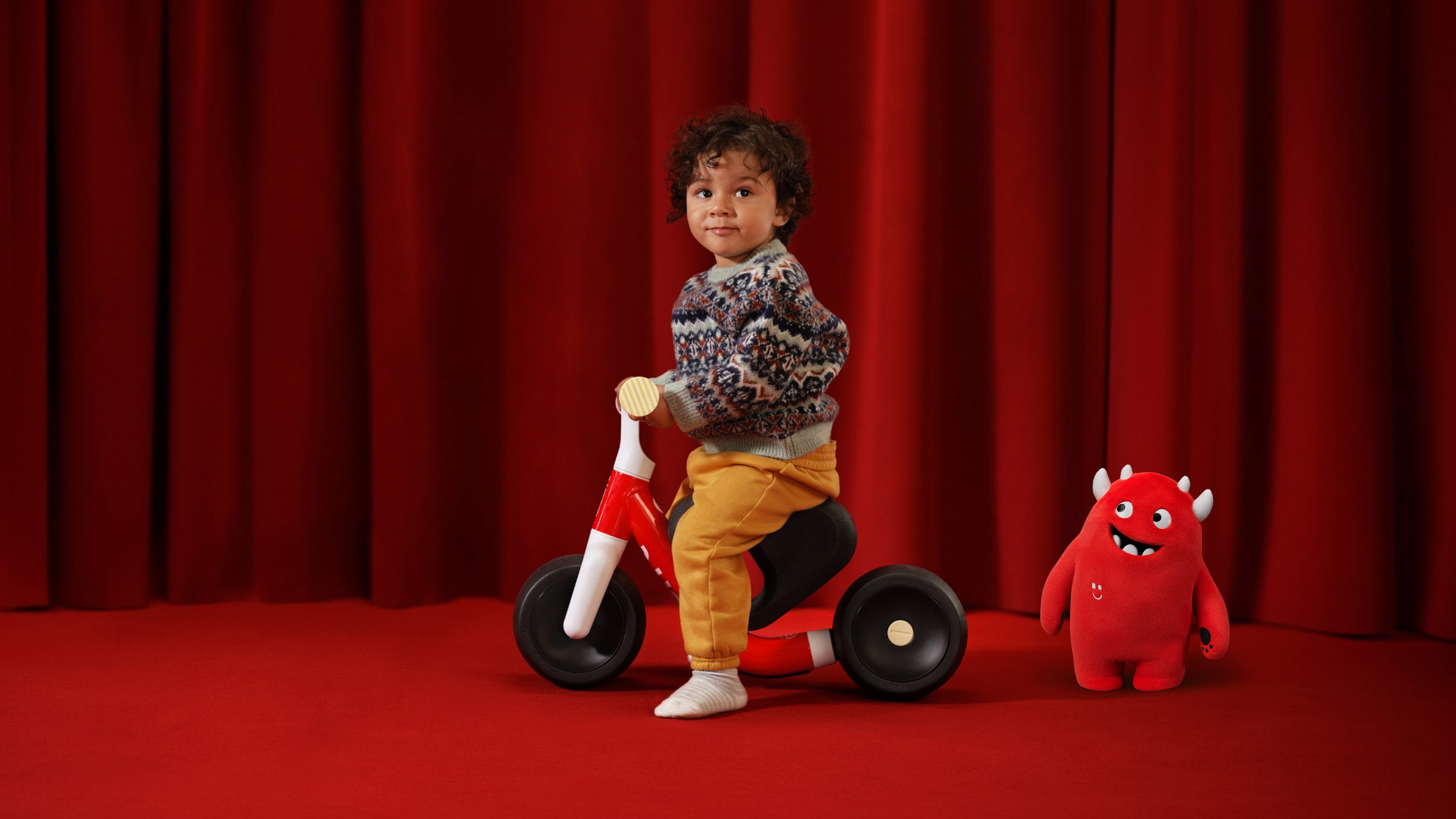 A happy toddler is sitting on the woom WOW in the living room with nothing on their feet and holding onto the handlebars.