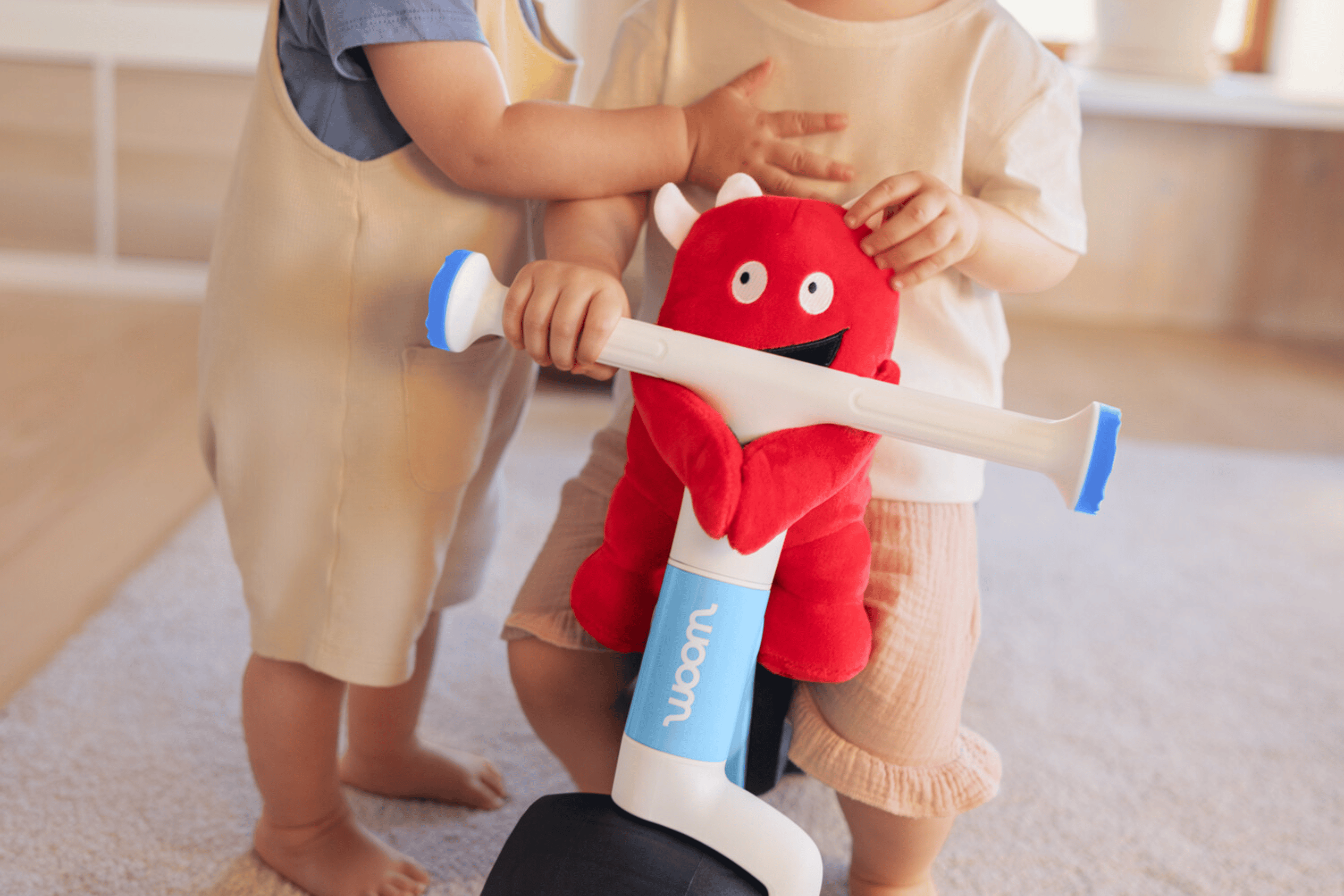 You can attach the soft toy to the handlebars on your child's woom WOW. A toddler is sitting on a bike, with another one next to him.