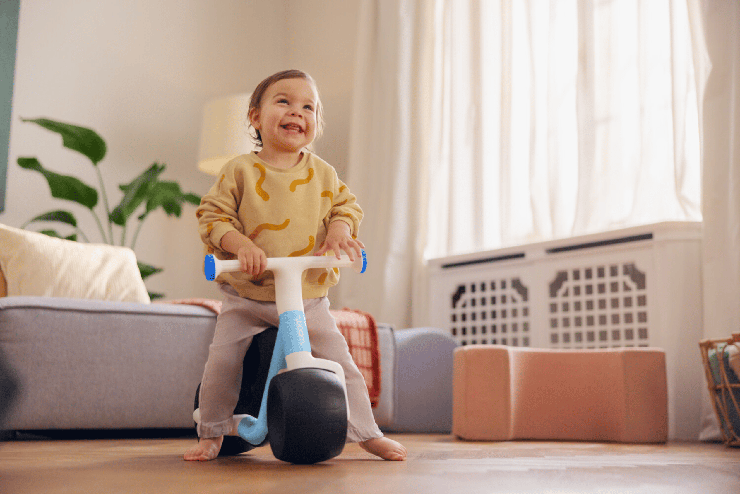 A happy toddler is sitting on the woom WOW in the living room with nothing on their feet and holding onto the handlebars.