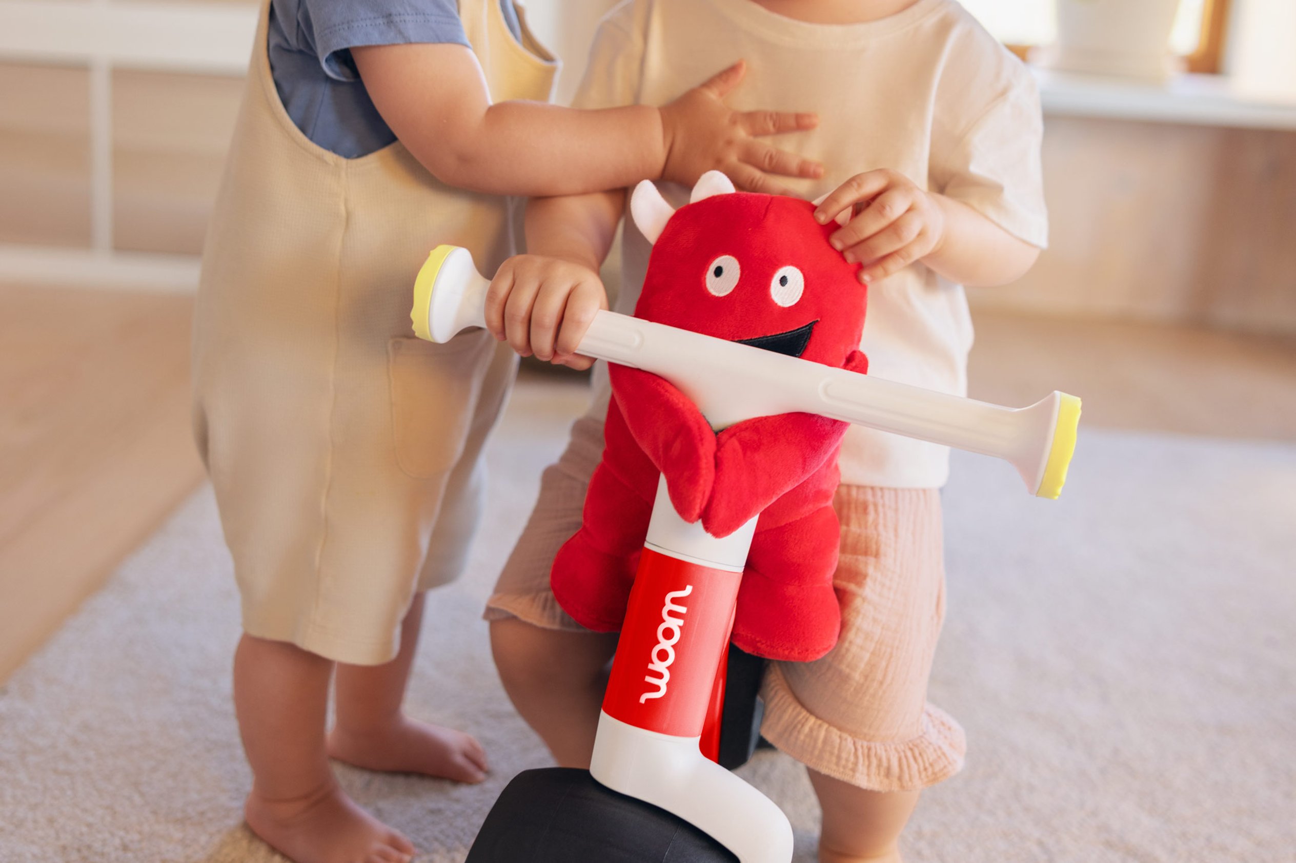 You can attach the soft toy to the handlebars on your child's woom WOW. A toddler is sitting on a bike, with another one next to him.