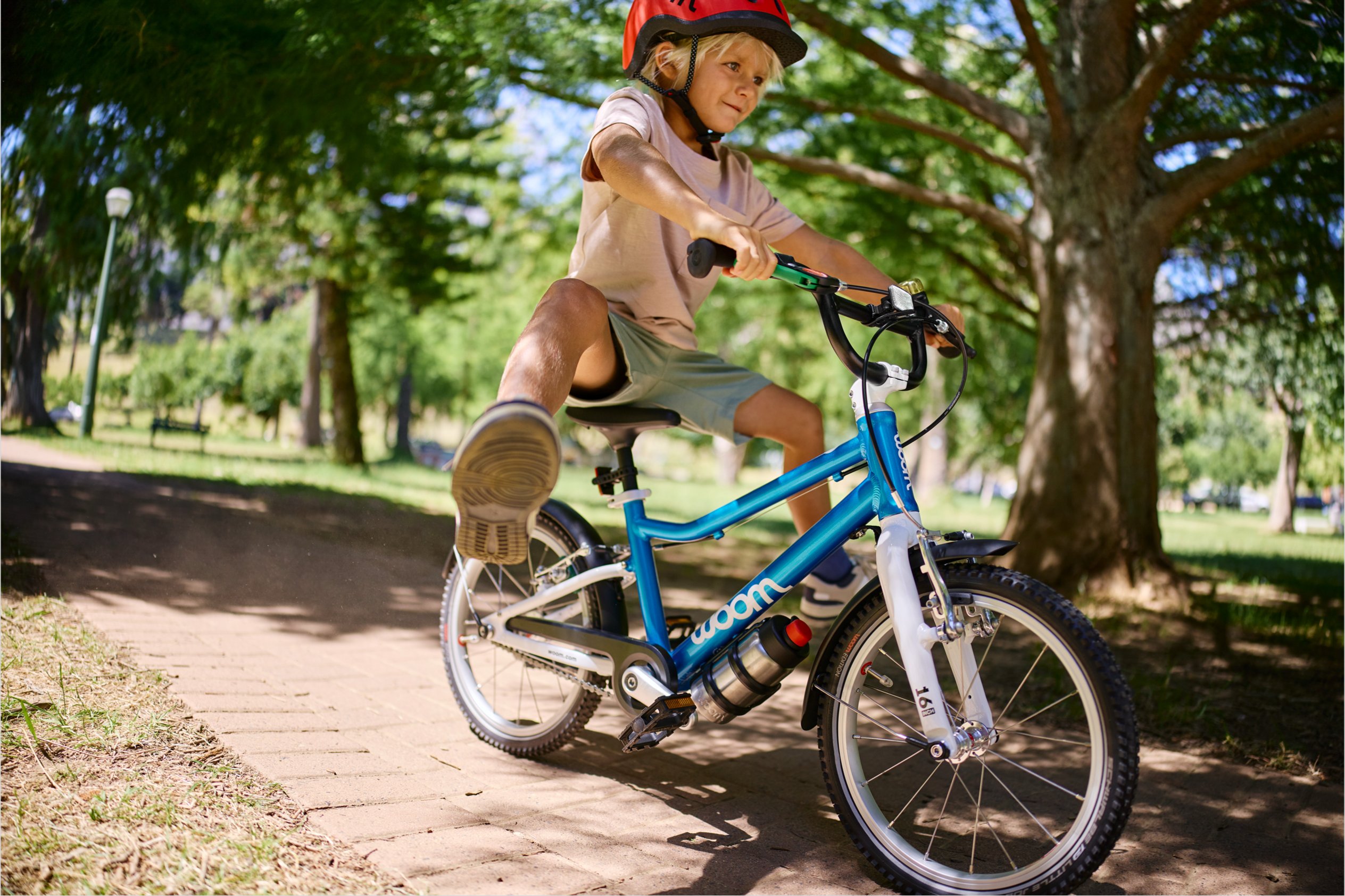 Junge mit rotem READY Kinderhelm fährt mit ausgestreckten Beinen auf dem Fahrrad woom GO 3 in Blau in einem Park.