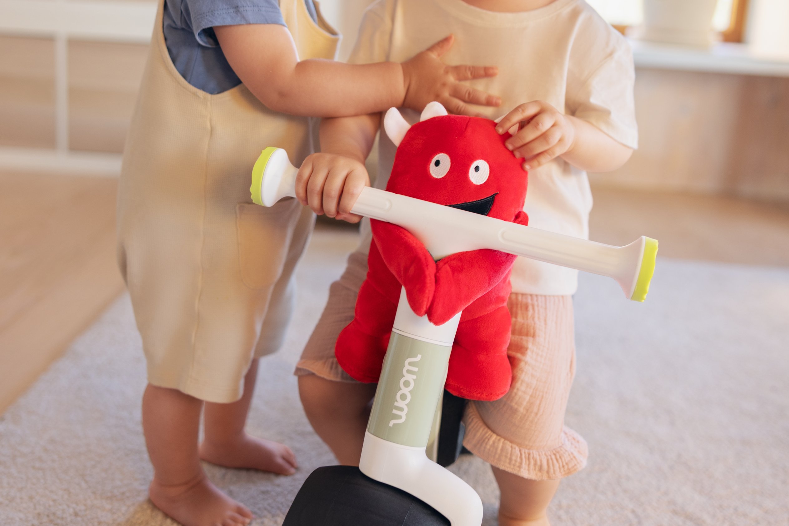 You can attach the soft toy to the handlebars on your child's woom WOW. A toddler is sitting on a bike, with another one next to him.