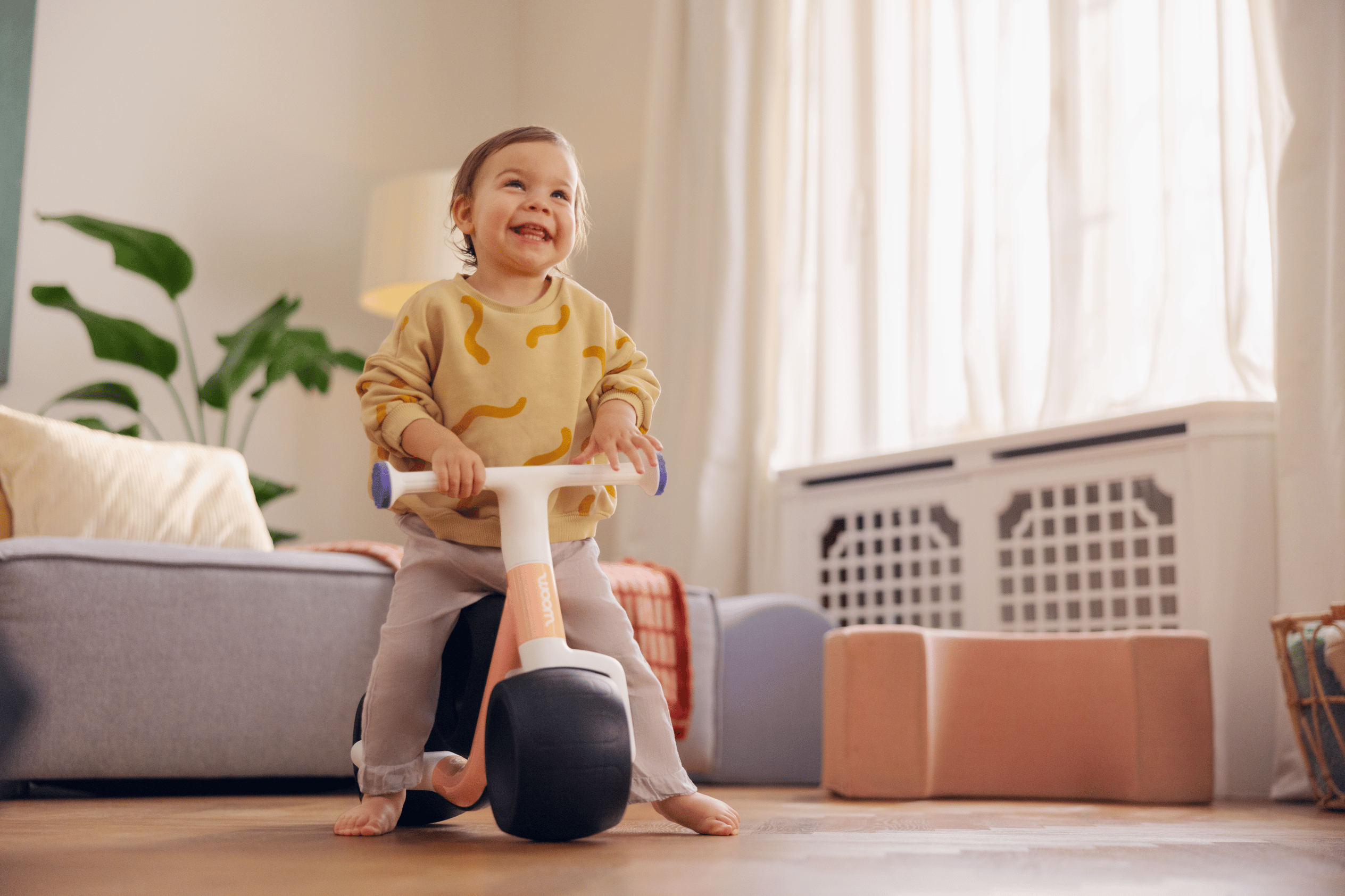A happy toddler is sitting on the woom WOW in the living room with nothing on their feet and holding onto the handlebars.