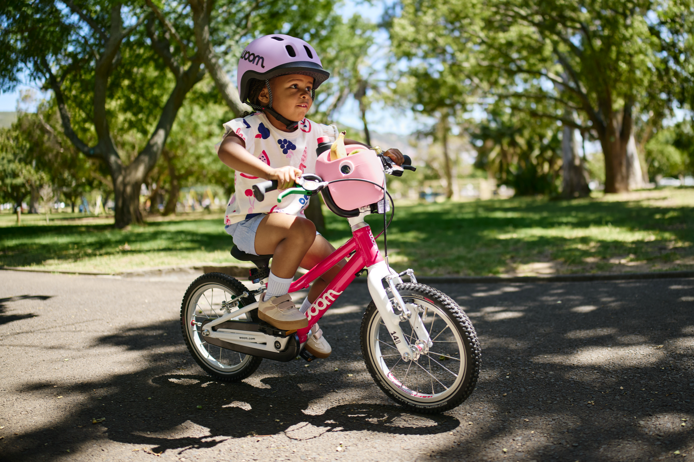 Kind mit READY Kinderhelm auf dem Kopf fährt auf dem Fahrrad woom GO 2 in der Farbe Pink. Auf dem Lenker steckt der POP Fahrradkorb in Pink.