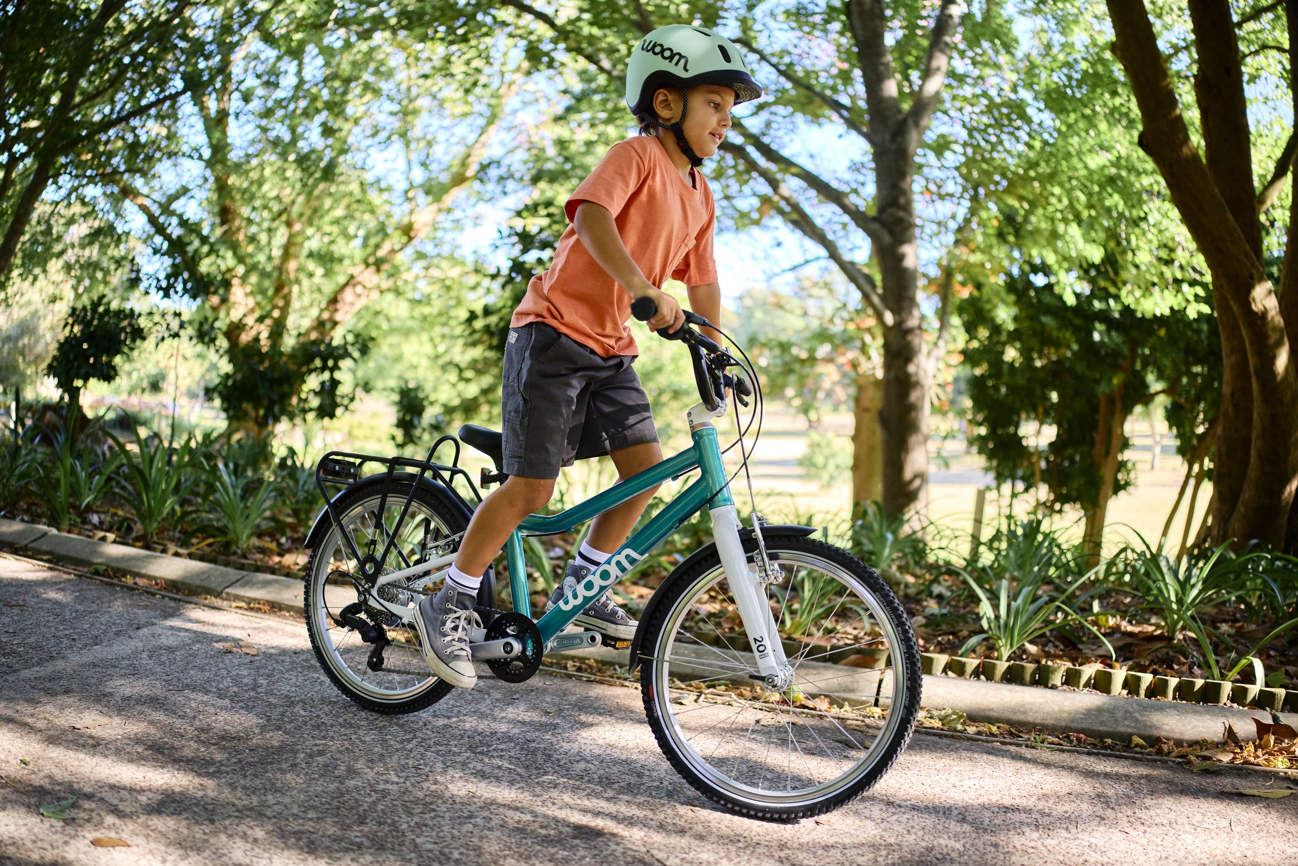 Junge mit grünem READY Kinderhelm auf dem Kopf fährt im Stehen auf dem woom GO 4 in der Farbe Minzgrün auf einem Weg im Grünen.