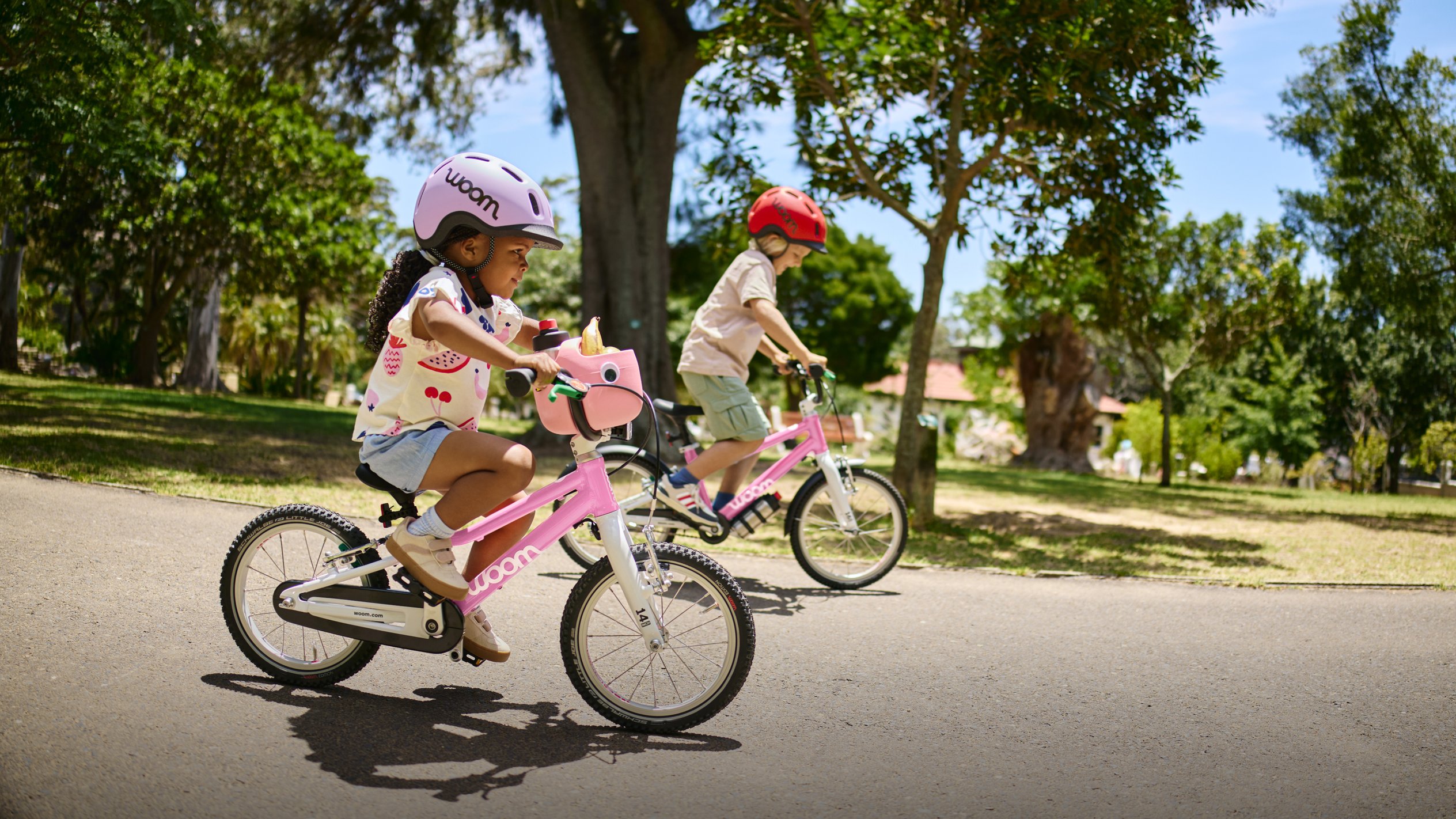 Junge mit rotem READY Kinderhelm fährt mit ausgestreckten Beinen auf dem Fahrrad woom GO 3 in Blau in einem Park.